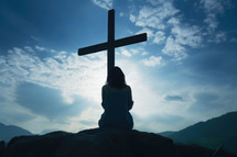 Young woman praying with a cross in the background at sundown