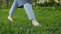 Female gardener in denim jeans and apron walking barefoot through lush green grass. Close up, slow motion view.