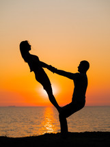  Silhouette of young woman and man doing pair yoga on sea beach at sunset. Meditation. Couple practicing acro yoga. Flexibility workout at nature background.
