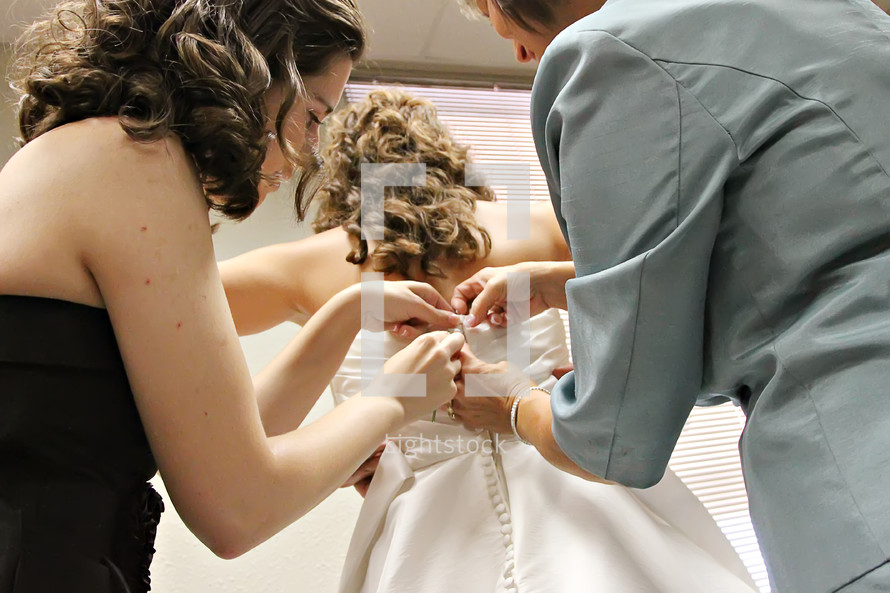Two women help a bride button her wedding dress.