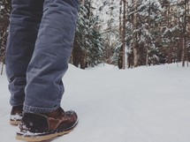 A man's legs and feet standing in a snow covered forest.