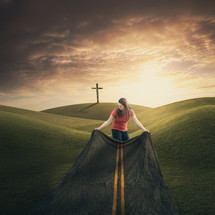Woman spreading a cloth on the ground that looks like a highway leading to a cross in the distance