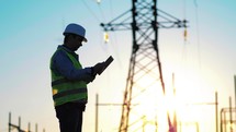 Power work concept. Engineer standing on field with electricity towers. Young engineer putting security helmet. Electrical engineer with high voltage electricity pylon at sunset background.