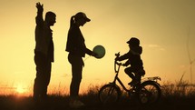 People silhouettes on summer sunset meadow. The kid is learning to ride a bike. Time to be together.Silhouette of happy family on a beautiful sunset meadow.
