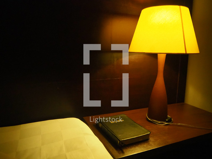 Bible on a wooden night table next to a bed with a lamp on it.