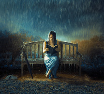 A woman sits on a bench in the rain alone while reading