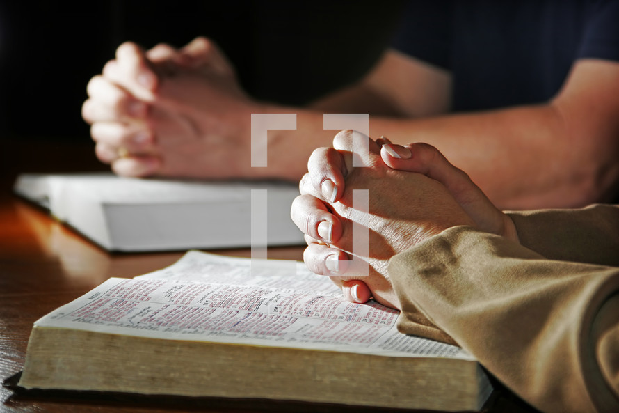 A woman and man fold their hands in prayer over their Holy Bibles - This can represent a devotion time for a couple or a couple being part of a larger bible study, prayer group, or small group from church.