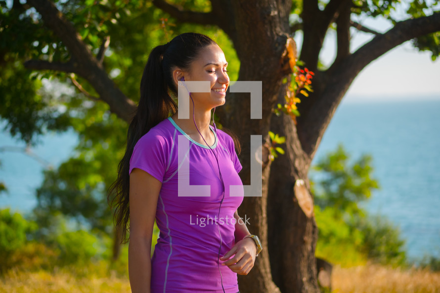 Portrait of young pretty sporty athletic woman listening to music with earphones on sea summer nature background. Lady after workout.
