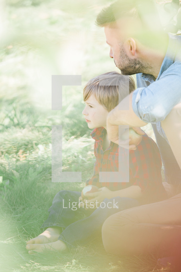 a father and son sitting outdoors holding an apple under bright sunlight 