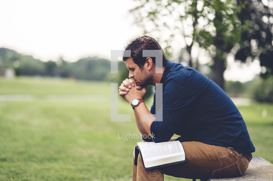a man praying sitting outdoors with a Bible on his lap 