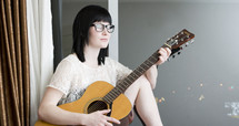Guitarrista; hipster woman with guitar and glasses sitting by a window.