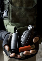 Man in military garb holding flashbang, fragmentation grenade, and shotgun cartridge.