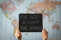 A woman in front of a world map holding a sign - For God so loved the world 