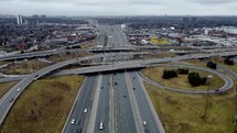 Aerial view of a drone flying over slightly busy Highway 401 in Toronto on a cloudy day. The drone is approaching a road interchange with moderate traffic speeding. Slight tilt downward of camera.