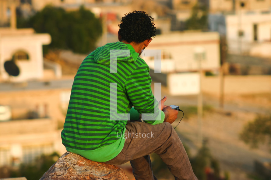 a man listening with earbuds sitting on a rock on a mountain 