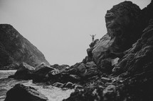 Woman standing on rocky cliff overlooking the ocean waves with arms raised.