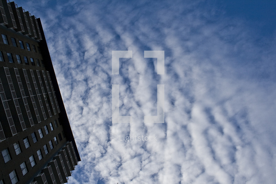 Ground view of side of tall building with cloud formations and blue sky in the background.
