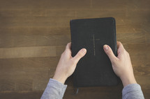 Hands holding a Bible on a wooden table.