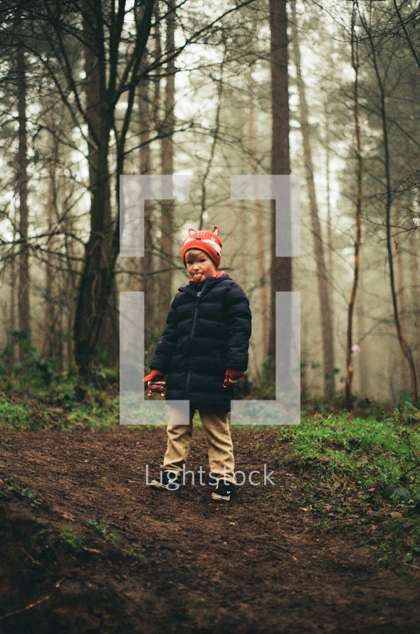 a boy in a coat standing in a forest 
