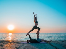  Young beautiful couple practicing acro yoga on the sea beach near water. Man and woman doing everyday practice outdoor on nature background. Healthy lifestyle concept.