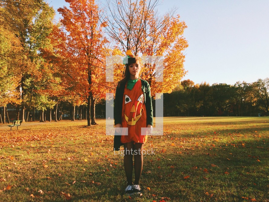 Girl in a pumpkin costume standing outside with fall foliage.