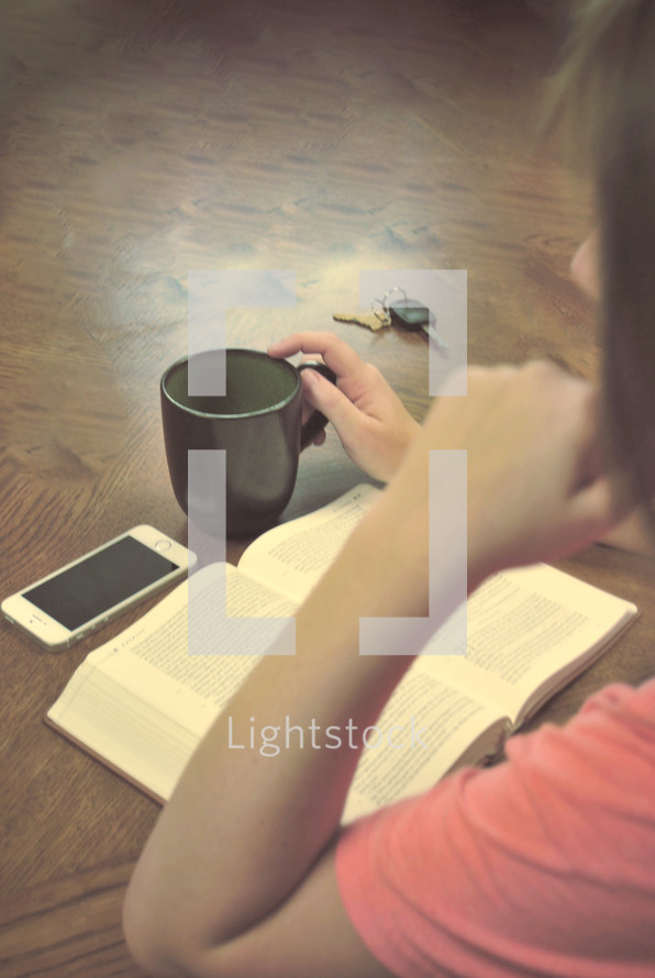woman reading a Bible with a coffee mug, car keys, and cellphone on the table 