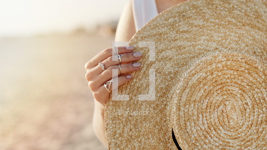 Woman hand with boho gypsy rings holding straw trendy hat. Girl standing on sandy beach at summer near sea and rolling her boater. High quality photo