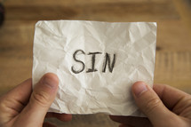 hands holding a piece of crumpled paper with the word sin.
