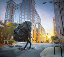A man struggles to walk across the street with a heavy stone he is carrying.