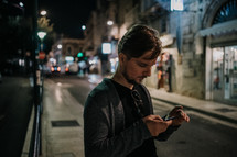 a man checking his cellphone on a dark sidewalk at night  in a city 