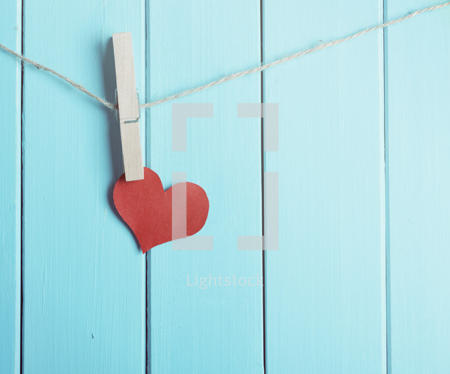 Clothespin holds red paper heart against a blue wall.