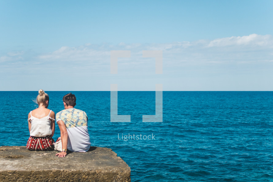couple sitting on a concrete wall looking out at the ocean 