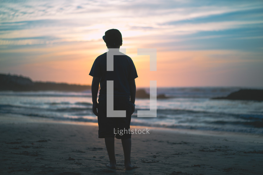 Silhouette of a young man standing on a beach at dusk | Youth | Facing Forward  | Praying | Sun | Faith | Children | Clouds | Courage | Determination | Mediation | Nature | Landscape | Peace