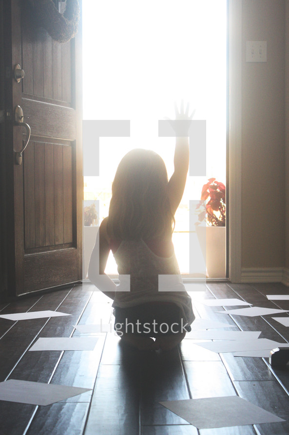 Silhouette of  a girl waving goodbye in a sunlit doorway surrounded by papers on the wood floor.