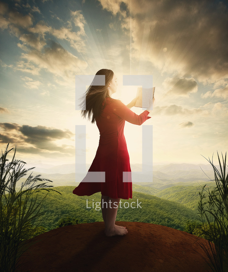 Woman standing on a hill reading the Bible with rays of light beaming from the clouds.