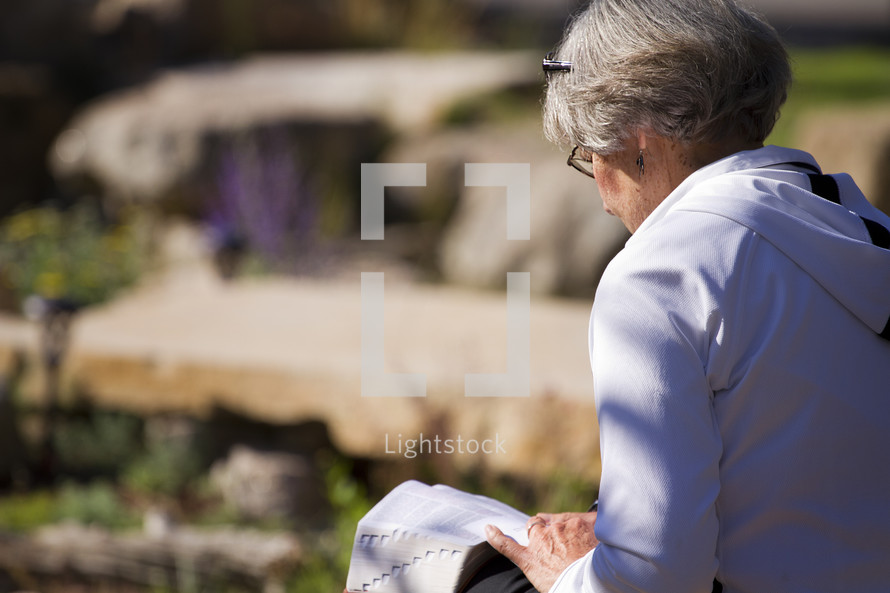 an elderly woman reading a Bible in her lap outdoors 