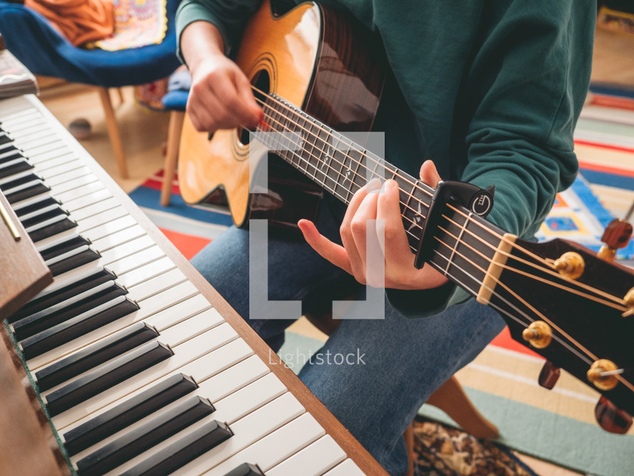a girl sitting at home in a living room  in front of a piano playing chords on an acoustic guitar