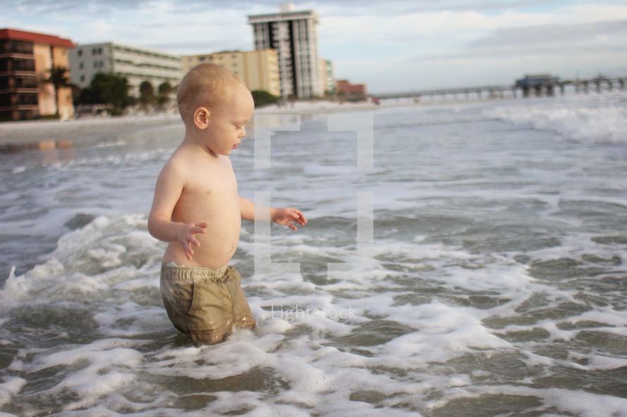 Boy walking in the ocean water near a pier and a hotel.