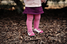Toddler girl's leg standing in wood chips with tree and sunset in background.