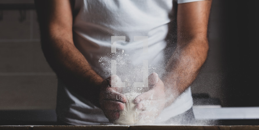 Man kneads raw dough, dust from flour flies in different directions, male hands on a black background