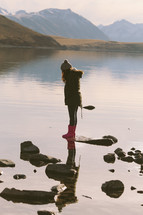 A girl standing on a rock at the edge of a lake in the mountains.