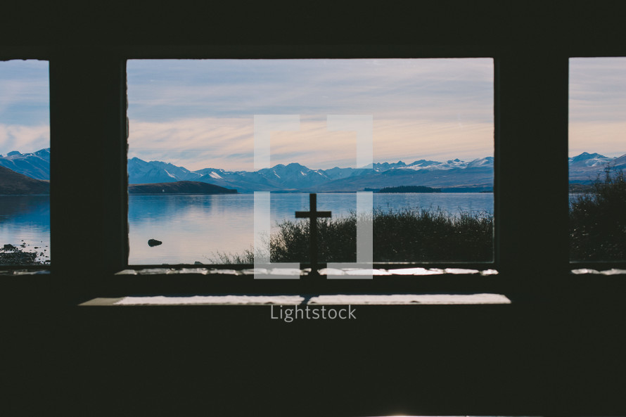 A cross on a windowsill looking out on a lake and mountain range.
