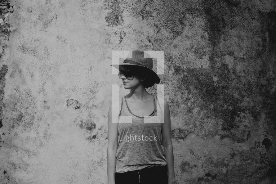 a woman in a hat and tank top standing in front of a grungy wall 
