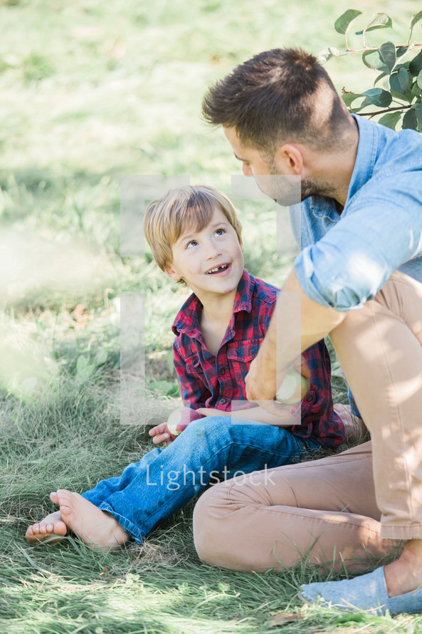 a father and son sitting in the grass holding apples 