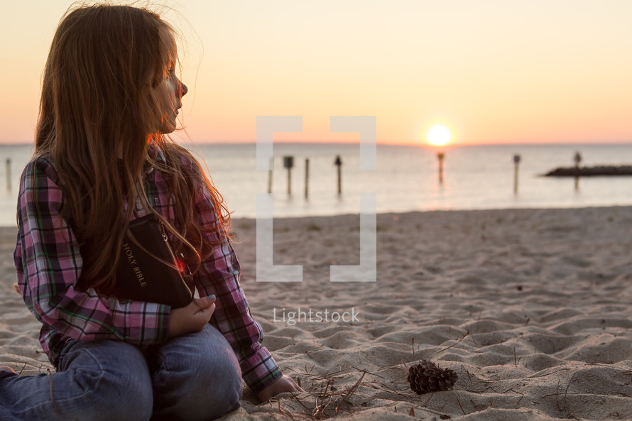 young girl sitting in the sand on the beach holding her bible at sunset