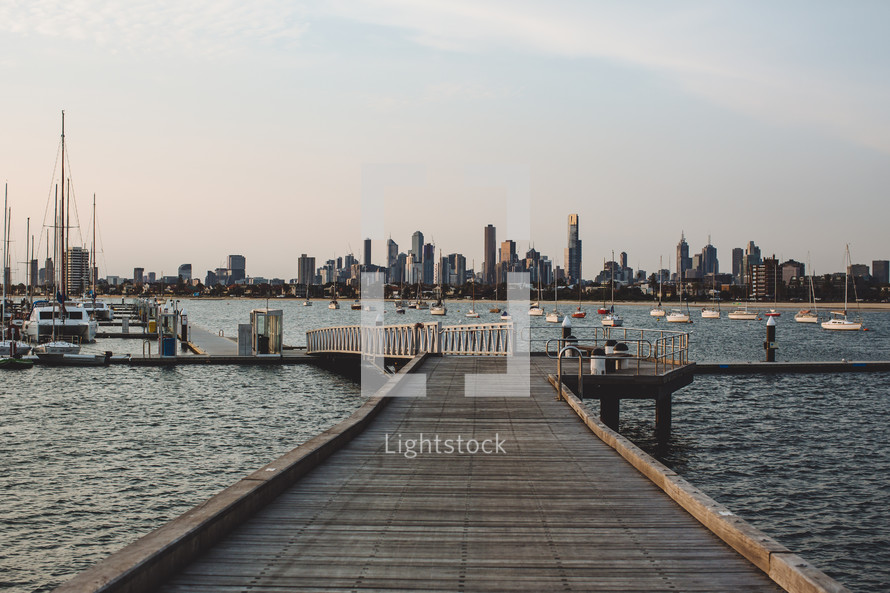 A wooden pier leading into a bay full of anchored sailboats with a city skyline in the background.