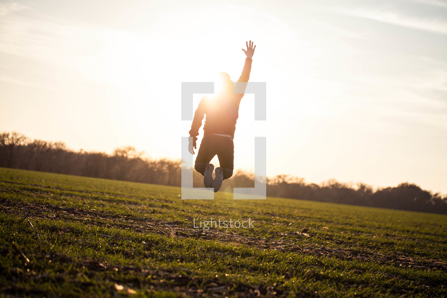 man jumping in a field with his hand raised to God under the glow of sunlight