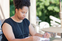 an African American woman sitting at a table outdoors reading a Bible 