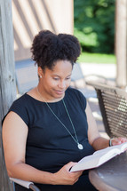 an African American woman sitting at a table outdoors reading a Bible 