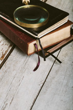 Magnifying glass on top of stack of massls and Bible on wooden floor.